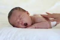 Yawning baby lying on their tummy, with a parents hand on their back.