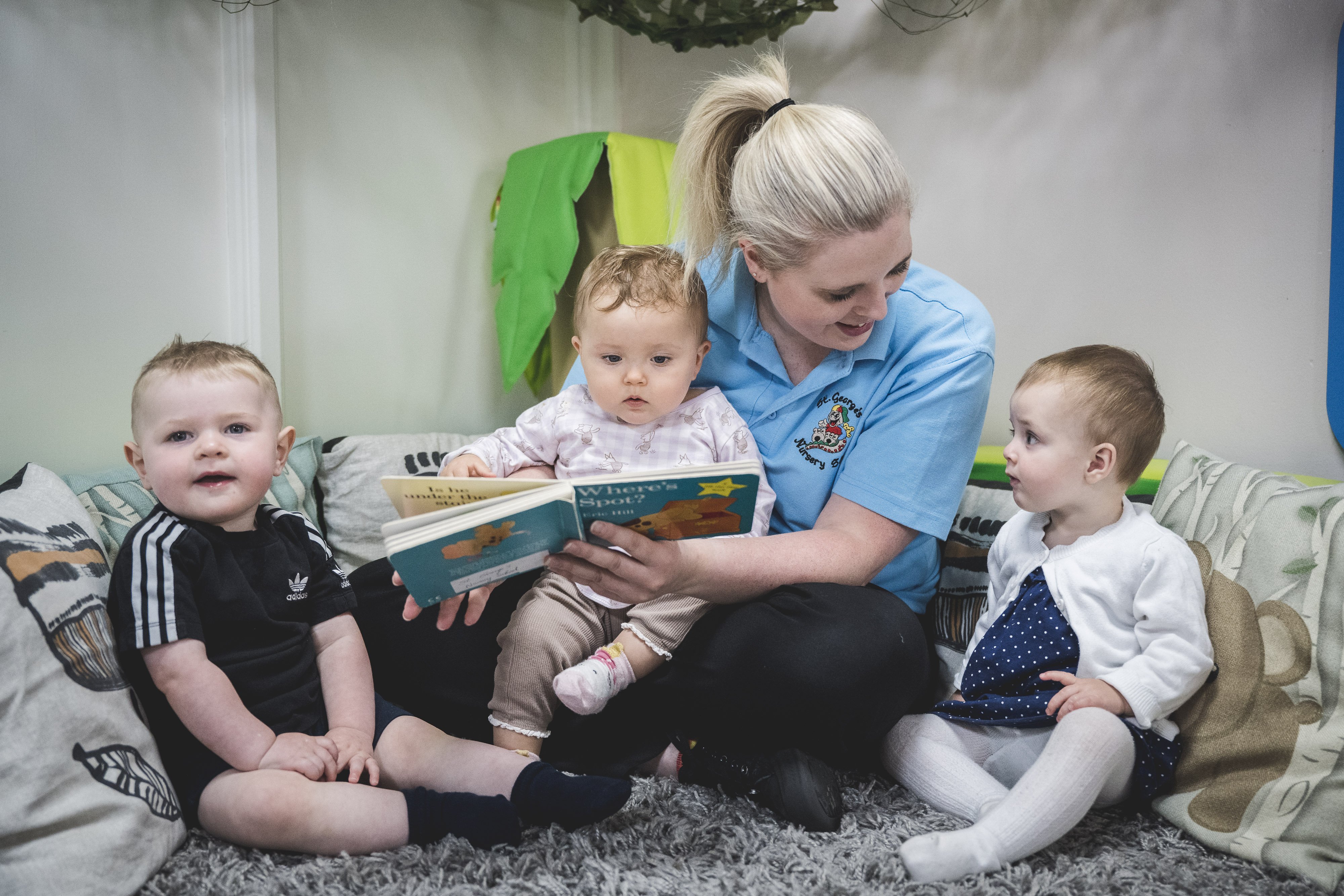 3 children sat with a nursery working on a rug with cushions.  Nursery worker reading Where's Spot? boardbook