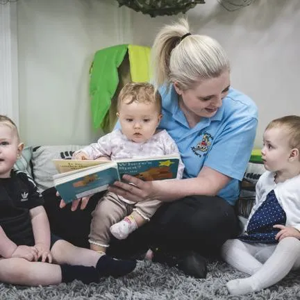 3 children sat with a nursery working on a rug with cushions. Nursery worker reading Where's Spot? boardbook