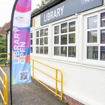 Woodthorpe Library exterior - View of an access ramp leading to a white one story building, windows along the side looking onto the shelves and a multicoloured Library Open flag at the foot of the ramp.